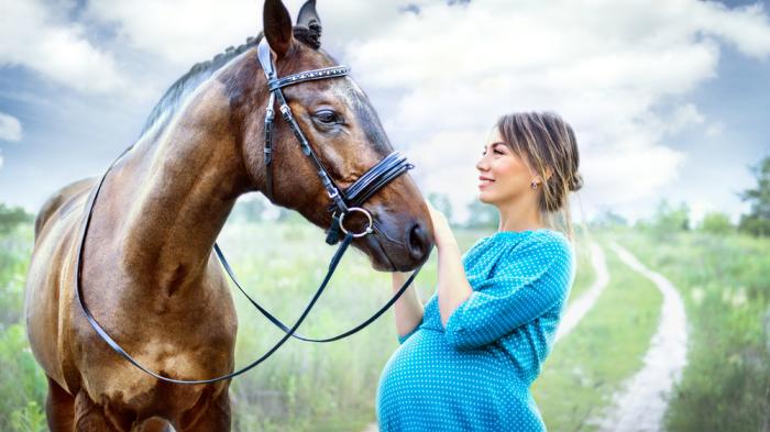 Braunes Pferd mit schwangerer Frau im blauen Kleid auf Feldweg