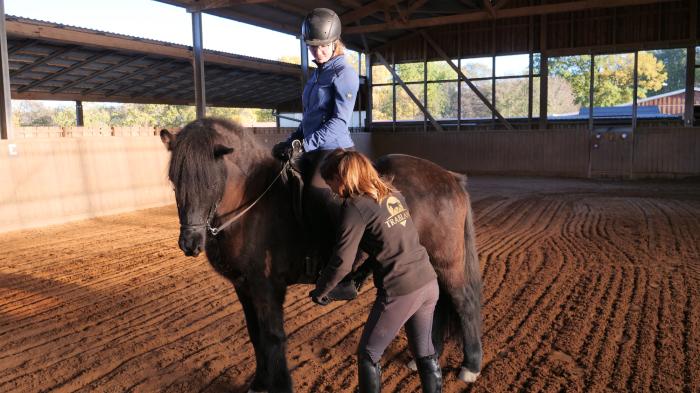 Eine Reitlehrerin hilft einer Frau mit blauer Jacke auf einem braunen Pony sitzend in einer Reithalle in den Steigbügel