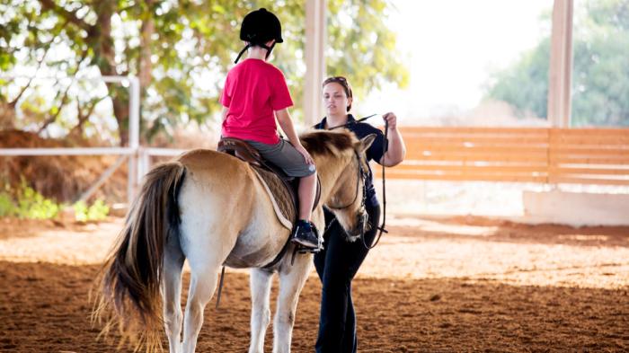 Eine Kind mit rotem Obertreil sitzt auf einem hellbraunen Pony in einer Reithalle, während eine Frau vor dem Pony steht und die Zügel um den Hals des Pferdes legt 