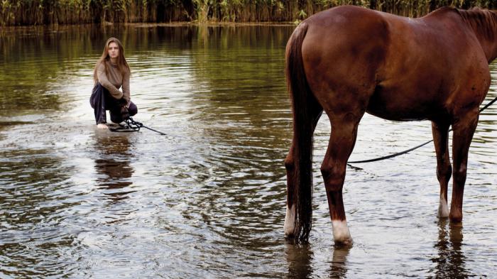 Eine Frau mit langen braunen Haaren kniet im flachen Wasser, während ein braunes Pferd, dessen Kopf man nicht sieht, neben ihr im See steht.