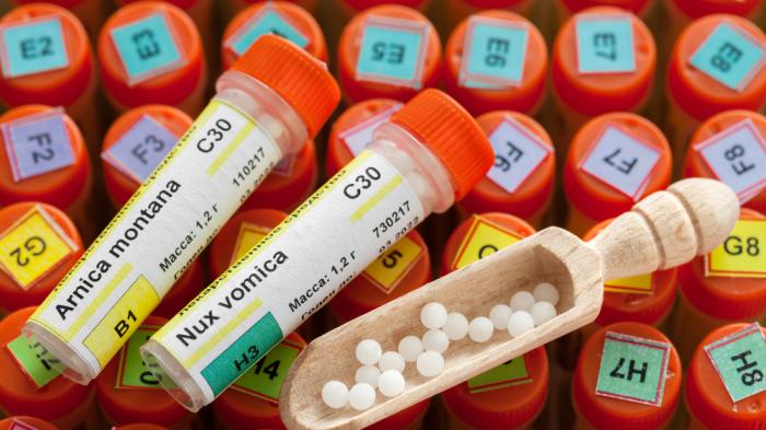 Nahaufnahme von Homöopathie-Globuli mit Beschriftungen "Arnica montana" und "Nux vomica". Im Vordergrund liegt ein Holzlöffel mit Globuli, im Hintergrund weitere bunte Röhrchen.
