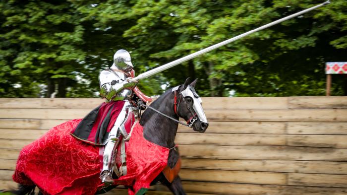 Ein Ritter in glänzender Rüstung reitet auf einem schwarzem Pferd, das mit einem roten Stoff bedeckt ist. Der Ritter hält eine lange Lanze und befindet sich in einer Turnierarena, umgeben von einem Holzzaun und Bäumen im Hintergrund.
