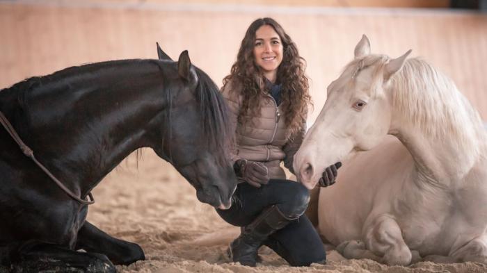 Eine Frau mit braunen, langen und lockigen Haaren kniet zwischen einem schwarzen und einem weißen Pferd, die auf dem Sandboden einer Reithalle liegen.