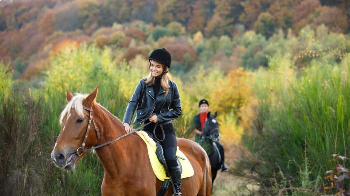 Zwei Reiterinnen mit Reithelm in Herbstlandschaft, eine blonde Frau mit schwarzer Lederjacke vorne auf einem braunen Pferd, die andere mit einem roten Oberteil dahinter auf einem schwarzen Pferd, beide tragen Helme.