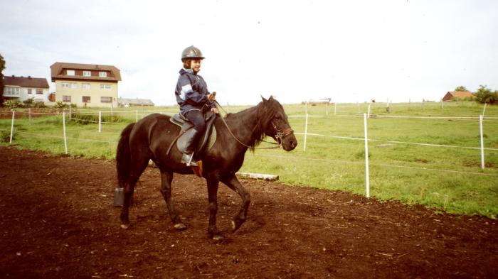 Ein Kind mit Helm und blauer Jacke reitet auf einem dunklen Pferd in einem umzäunten Bereich. Im Hintergrund stehen Wohnhäuser und Wiesen.