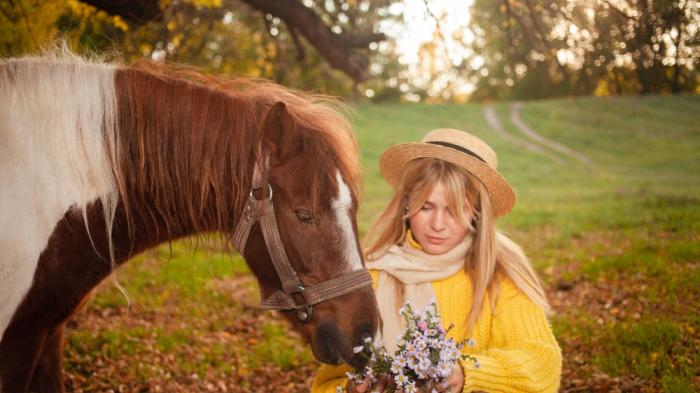 Frau mir gelben Oberteil und Strohhut füttert ein braun-weißes Pony mit Blumen