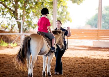 Kind sitzt auf Pferd beim Therapeutischen Reiten mit Therapeutin