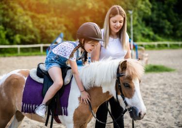 Kind auf Pony auf Reitplatz mit Teenager während Reiterferien für Kinder