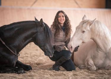 Eine Frau mit braunen, langen und lockigen Haaren kniet zwischen einem schwarzen und einem weißen Pferd, die auf dem Sandboden einer Reithalle liegen.